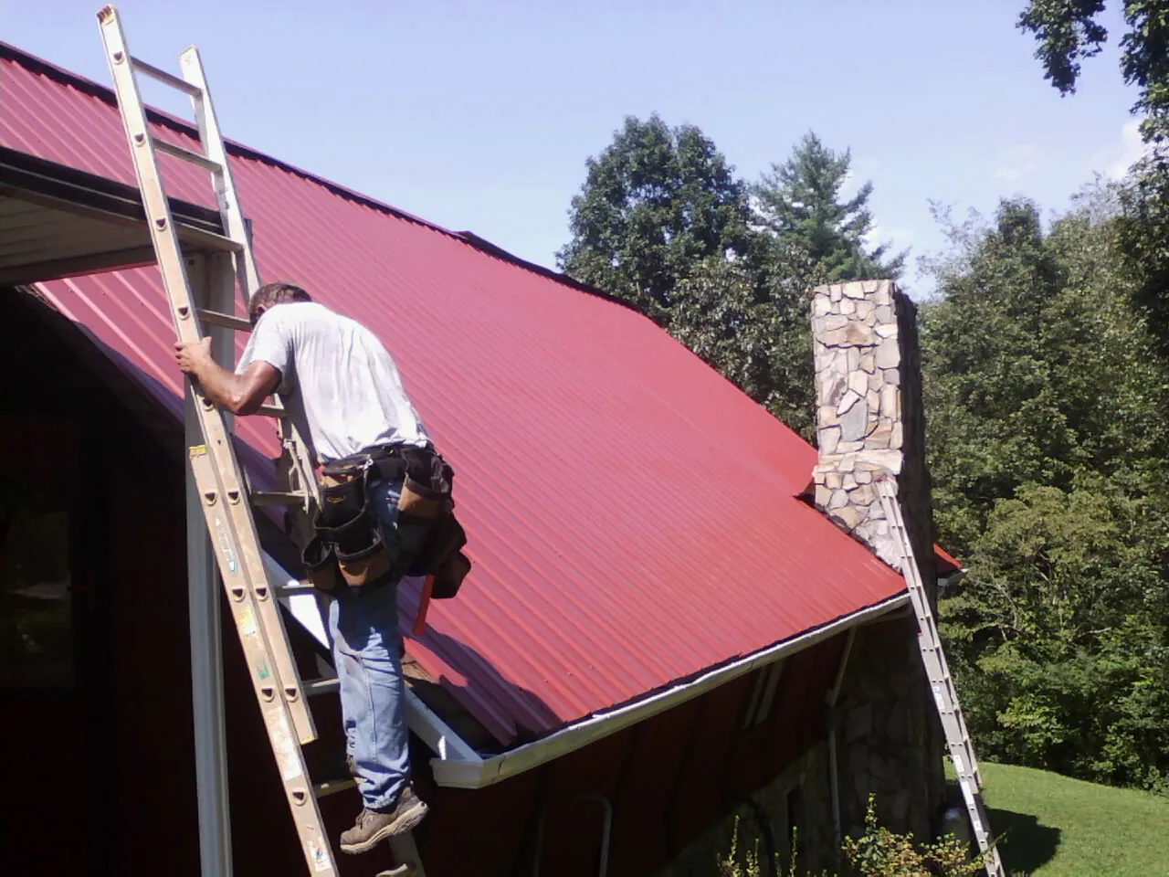 Roofer on a ladder beside a red metal roof and stone chimney — Soffit & Fascia Repair by Roofing Brothers in Concho